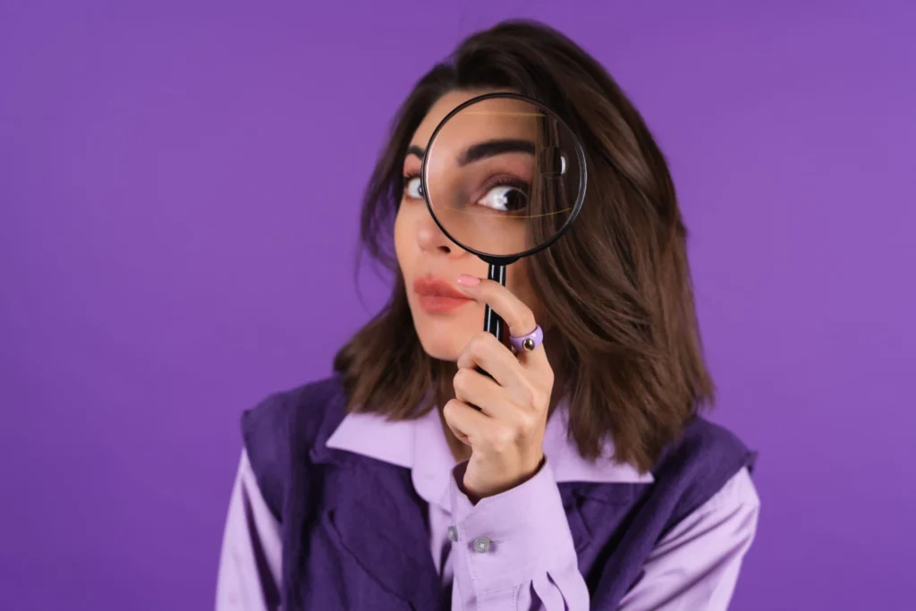 Foto de uma mulher em um fundo roxo com uma lupa na frente do olho, representando a pesquisa que um paciente realiza antes de agendar um psicólogo.